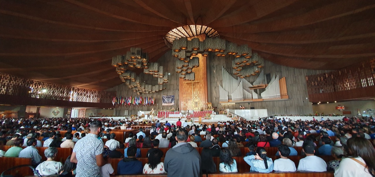 Mass in the modern Basilica of Guadalupe, the crowd filling the nave beneath the sweeping wooden roof, with the tilma above the high altar.