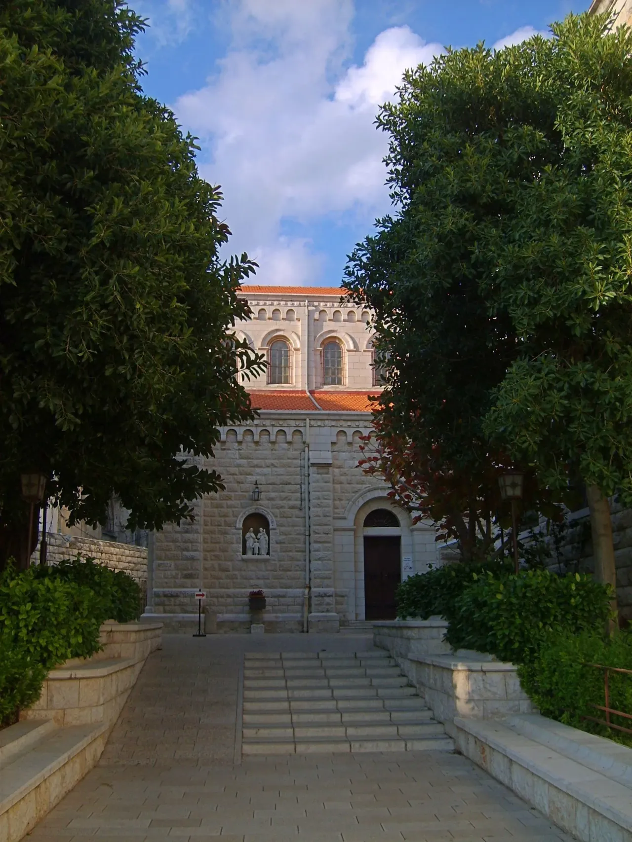 Steps and a stone path between trees lead to the door of Saint Joseph's Church in Nazareth, with a Holy Family statue in a niche beside it.