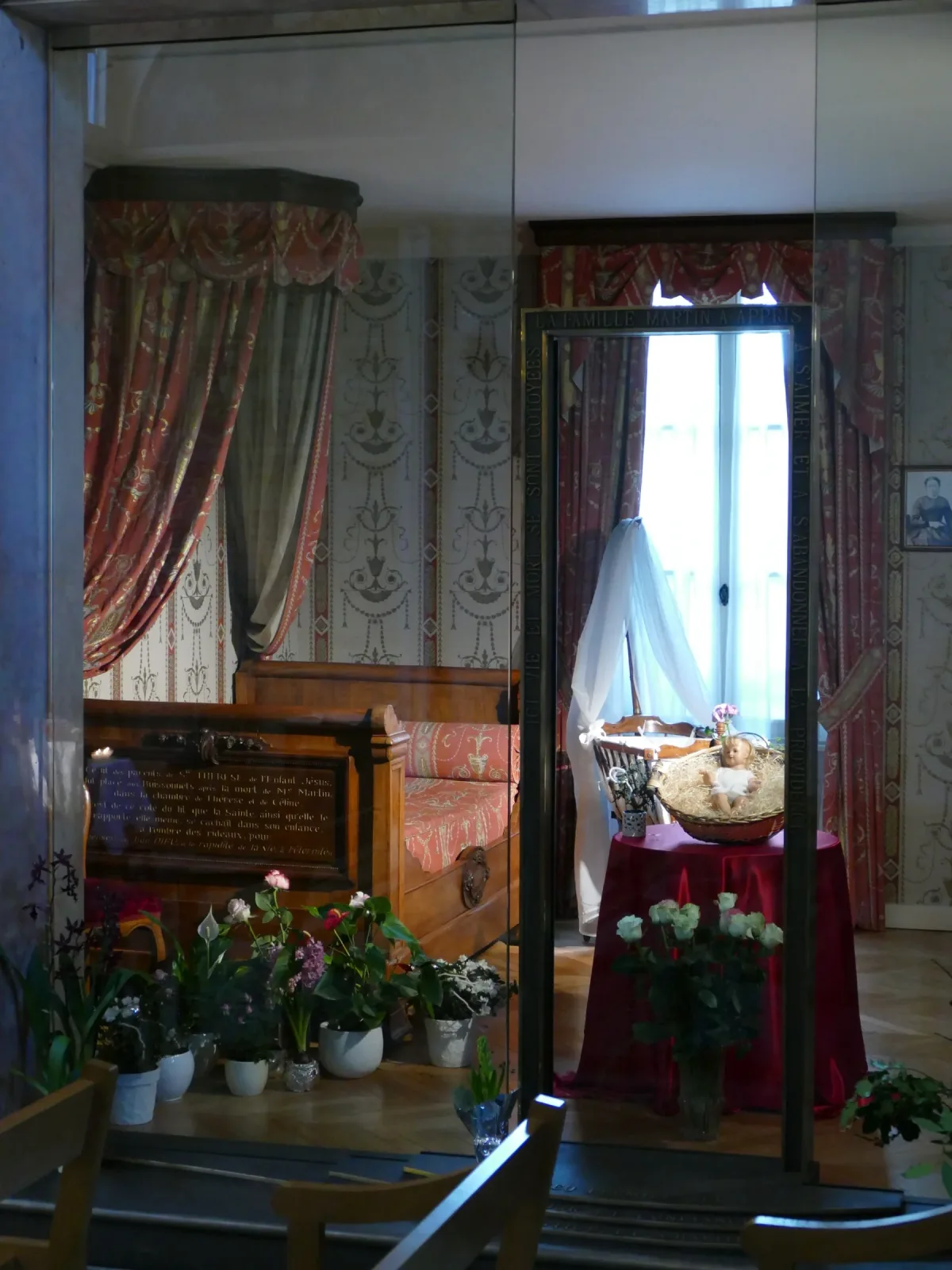 The birth room of Saint Thérèse at 50 rue Saint-Blaise in Alençon, seen through the glazed bronze doorway from the chapel, with a bed, a draped cradle and flowers left by pilgrims.