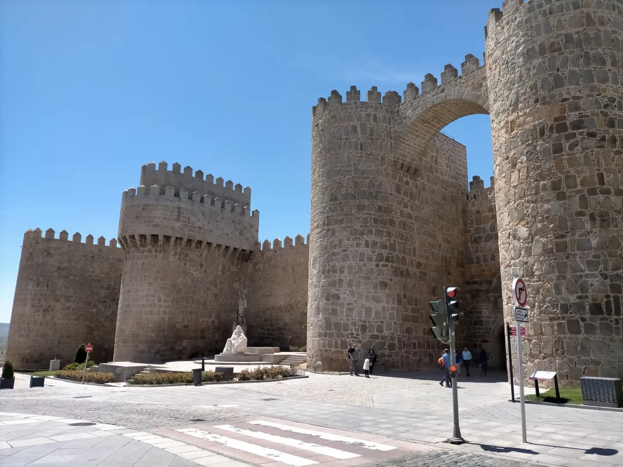 The Puerta del Alcazar in the walls of Avila, two round towers flanking the arch, with the seated monument to Saint Teresa on the pavement in front of them.