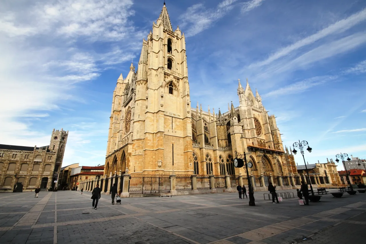 León Cathedral in morning light from the Plaza de Regla: the south tower of the west front with its spire, and the long south flank of buttresses and pinnacles under a high, streaked sky.