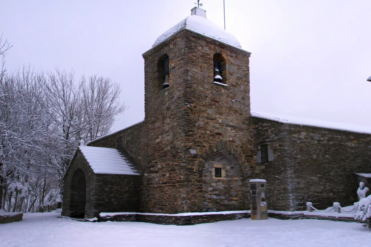Santa María a Real at O Cebreiro under snow and a pale overcast sky: the square stone bell tower with its two bells, the snow-covered roof of the porch, and frosted trees beside the church.