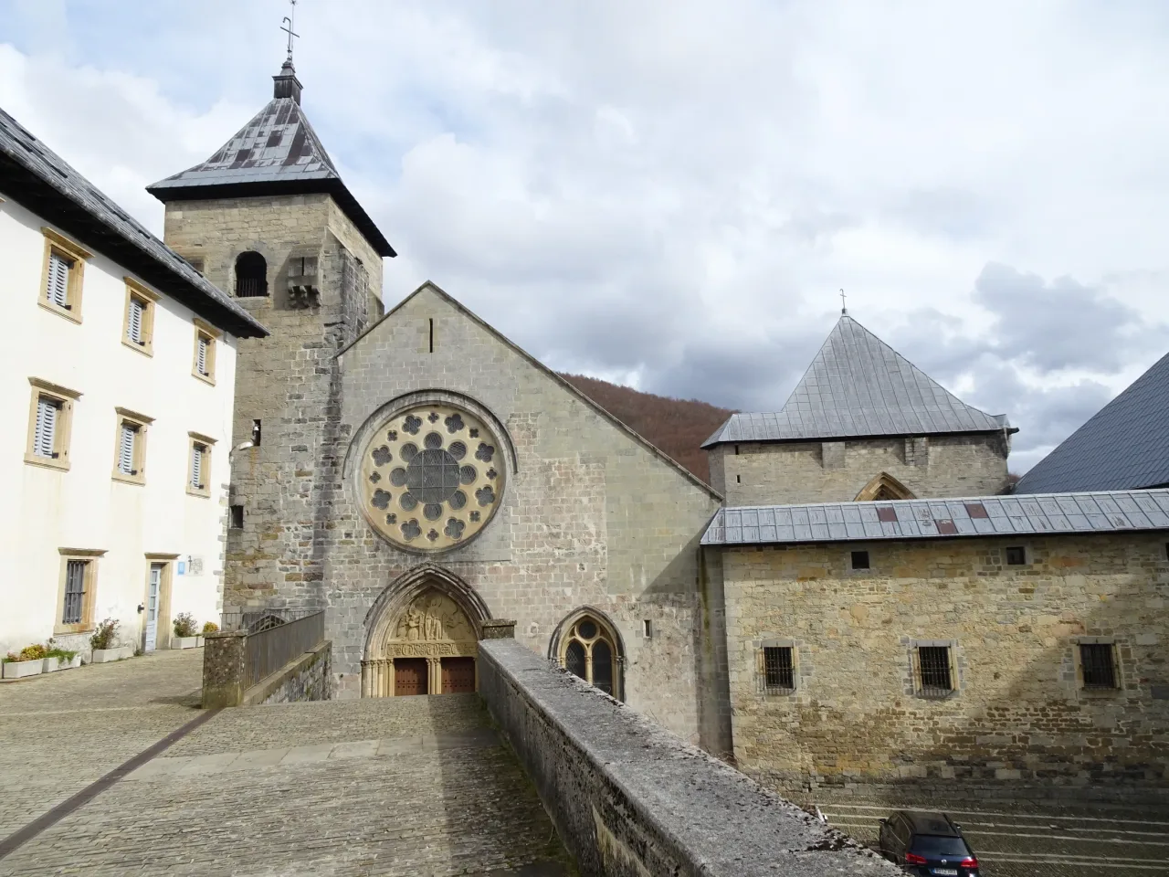 The stone front of the collegiate church at Roncesvalles under a soft overcast sky: the rose window above the carved portal, the bell tower, and the square chapel of San Agustín beyond, with a ramp leading up to the door.