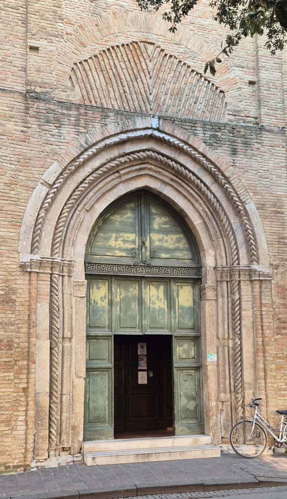 The pointed medieval portal of the basilica in Forlì, its twisted stone colonnettes set into a plain brick wall, one green door standing open.