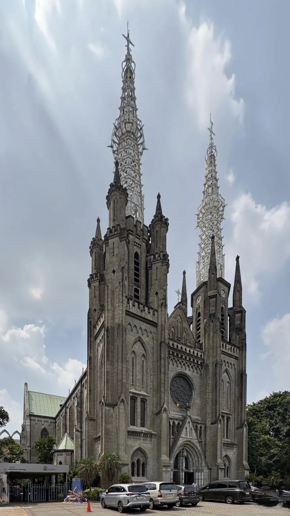 The neo-Gothic facade and twin spires of Jakarta Cathedral, with the rose window and parked cars on the forecourt.