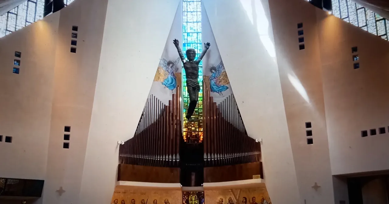 The sanctuary wall of the Church of the Redeemer at Materdomini, a bronze risen Christ with arms raised against a tall stained-glass window above the organ pipes.