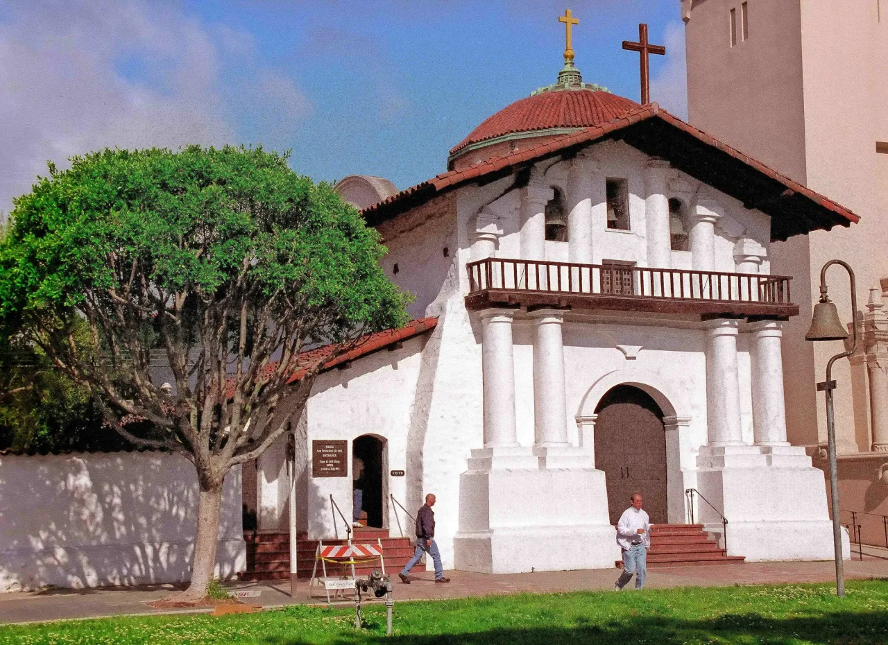 The white adobe front of the Old Mission chapel at Mission Dolores, with its columned porch, balcony and three bells.
