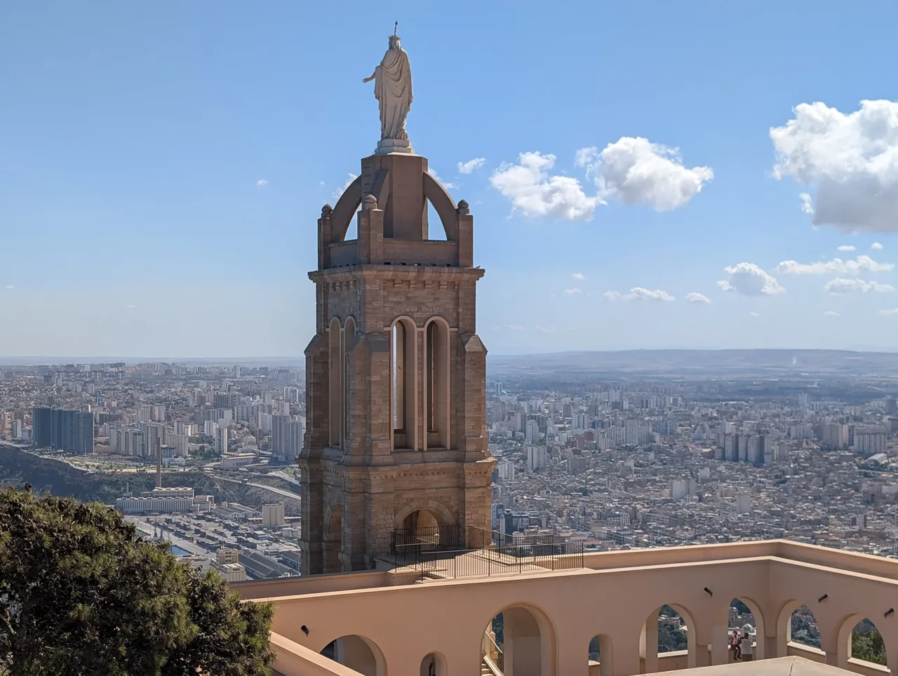 The tower of the sanctuary of Notre-Dame de Santa Cruz crowned by the statue of the Virgin, with the arcaded esplanade below and the city of Oran spread out beyond.