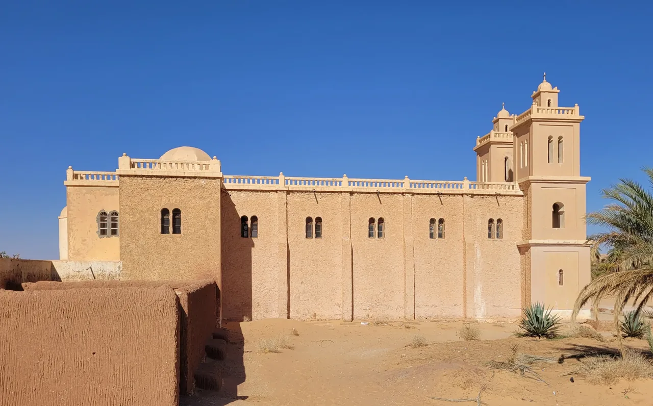 The ochre mudbrick church of Saint Joseph at El Menia, with its tower and a palm against a deep blue Saharan sky.
