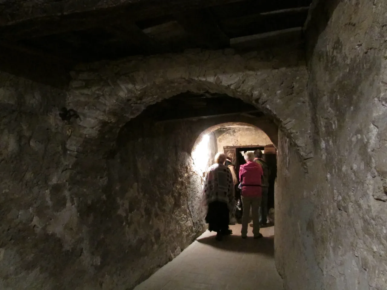A low stone passage of the first friary at Greccio, with pilgrims walking toward a lit doorway.