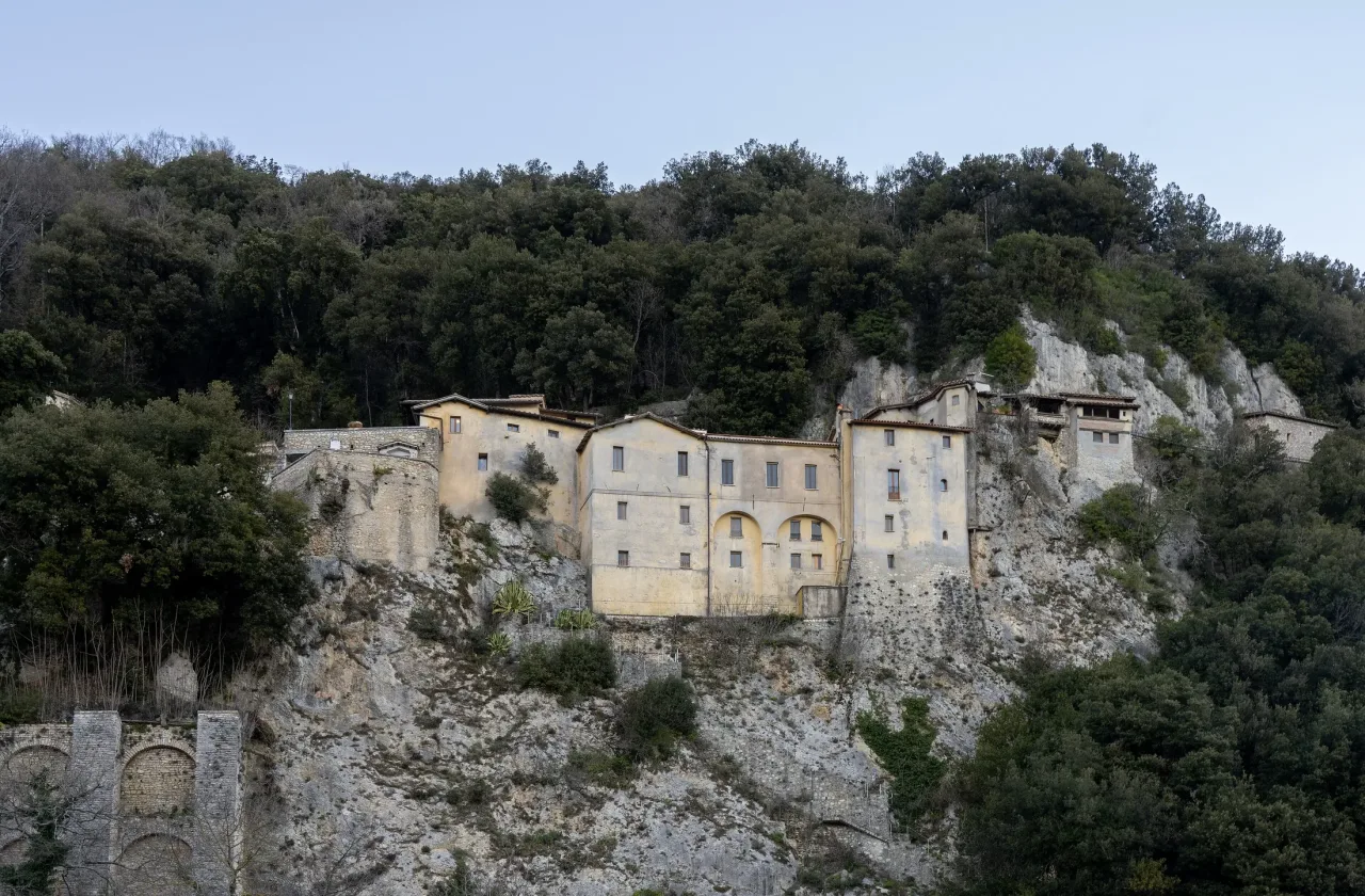 The pale stone buildings of the Greccio sanctuary built along a limestone cliff above dense oak woods.