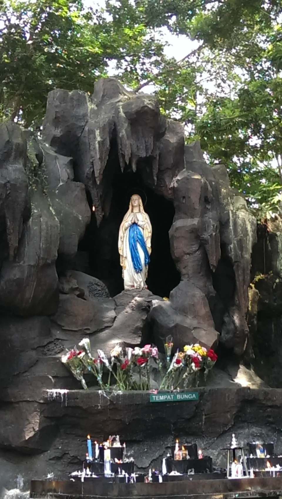 The Lourdes statue of Our Lady in the rock grotto at Kerep Ambarawa, with flowers on the ledge and candles below.