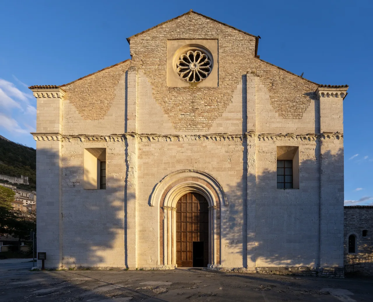 The plain limestone facade of San Francesco in Gubbio, with a round rose window and a pointed portal, in low evening light.