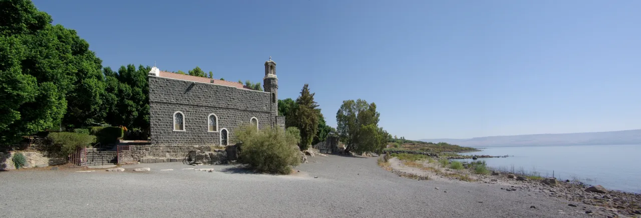 The dark basalt Chapel of the Primacy of Saint Peter beside a gravel shore path, with the Sea of Galilee opening to the right.