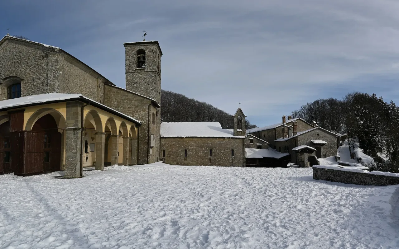 The basilica portico, bell tower and stone buildings of the La Verna sanctuary around the Quadrant square under snow, with fir forest behind.