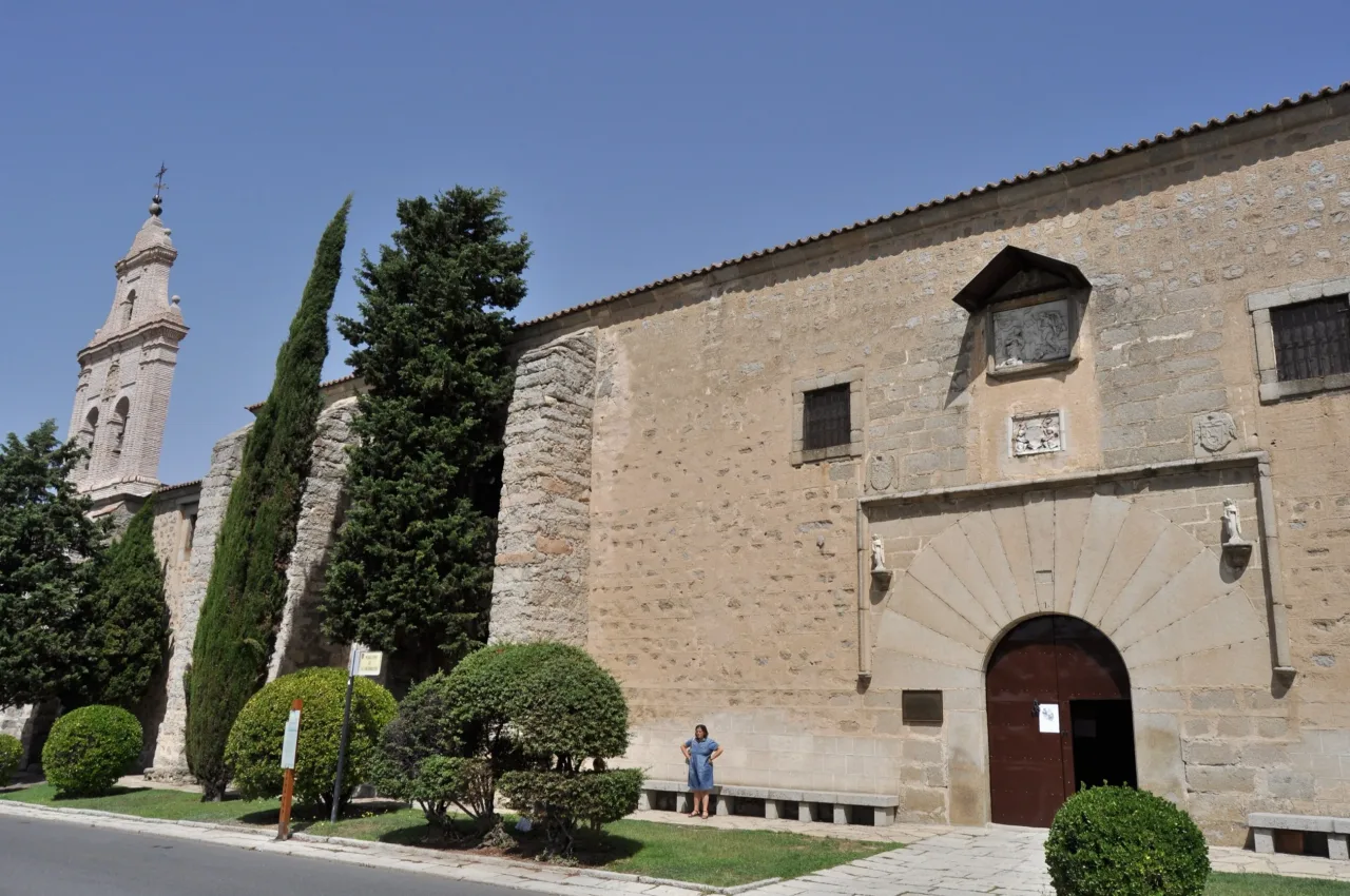 The long stone flank of the Monastery of the Incarnation in Avila, its doorway framed by a fan of granite voussoirs, with cypresses and the bell tower of the church beyond.