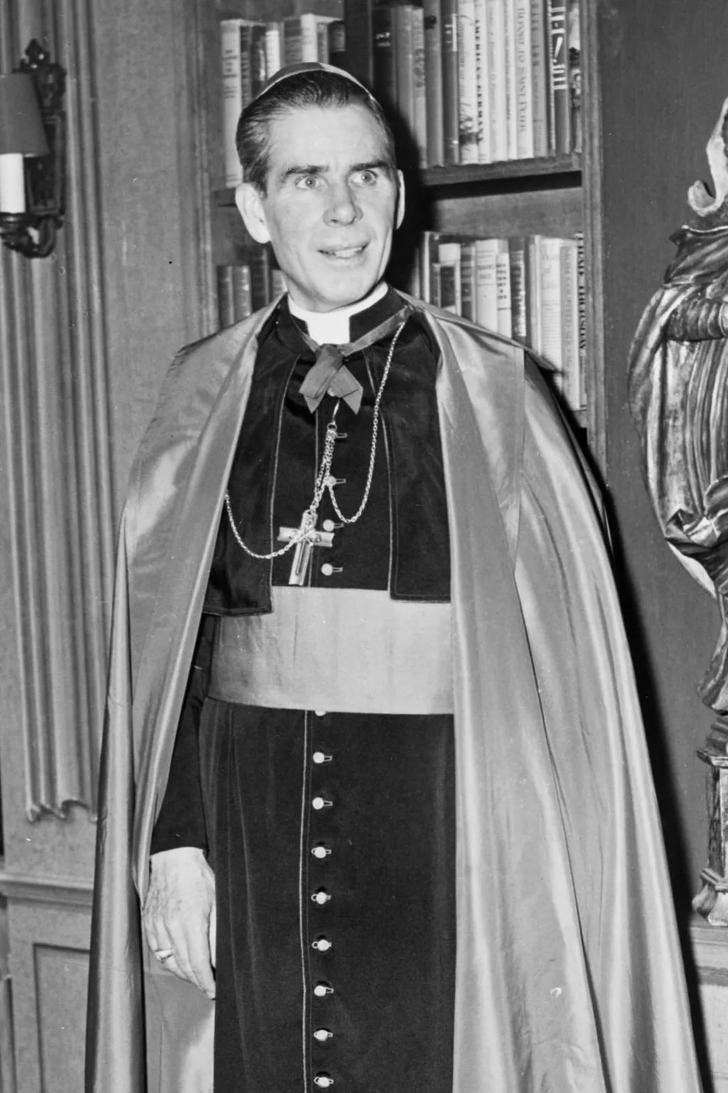 Fulton J. Sheen in episcopal cape with pectoral cross, standing in a library beside a carved statue of the Madonna and Child, photographed in 1952.