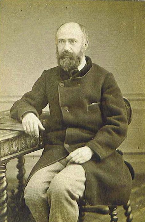Studio photograph of Louis Martin, bearded and in a dark buttoned coat, seated beside a carved table.